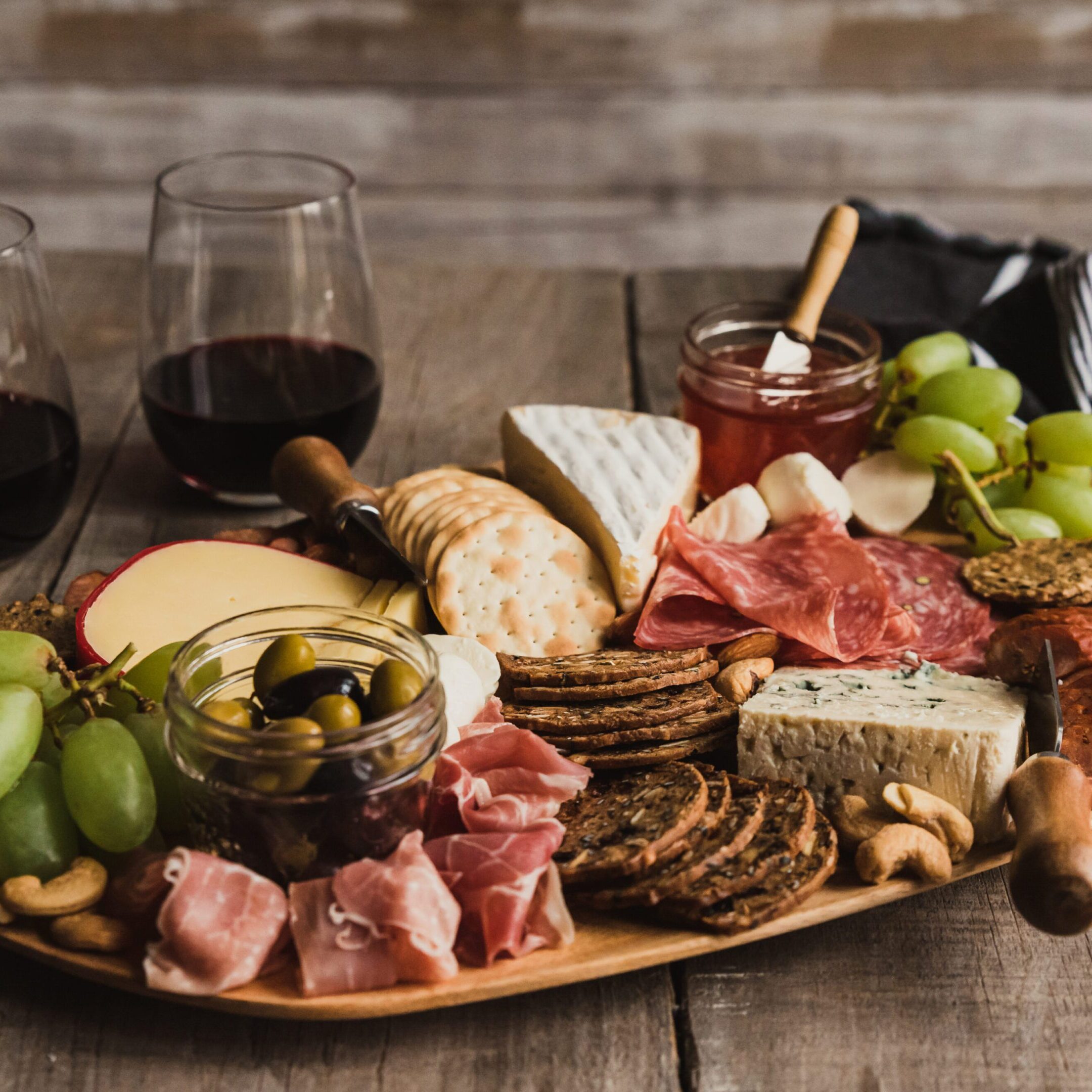 Close up of charcuterie board and glasses of wine on wooden table. A wooden platter with cheese, crackers and grapes.